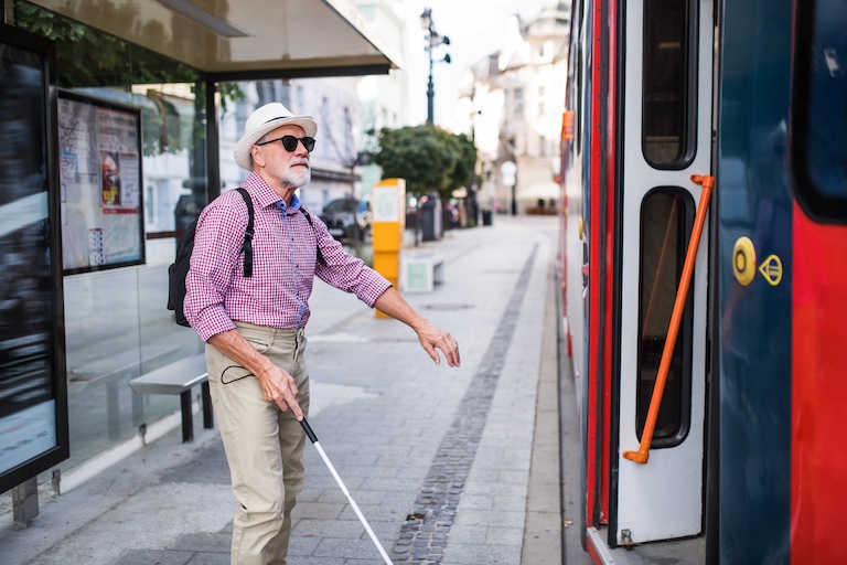 A senior blind man with white cane getting on public transport in city.
